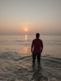 Rear view of man standing in sea against sky during sunset