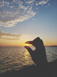 Silhouette person hand holding sea against sky during sunset