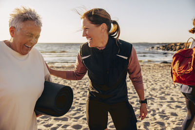 Happy female coach laughing with senior woman holding exercise mat at beach