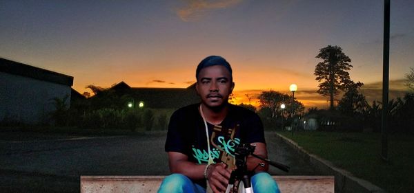 Portrait of young man sitting on road against sky during sunset