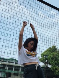 Portrait of young woman with arms raised standing against fence