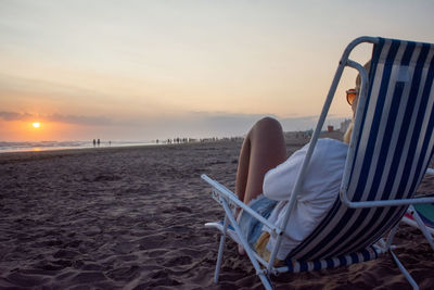 Man sitting on chair at beach against sky during sunset