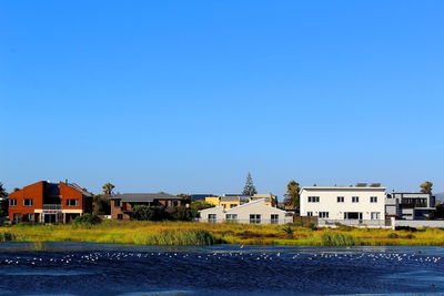 Houses by river and buildings against clear blue sky