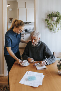 Female care assistant serving tea to senior man sitting at home