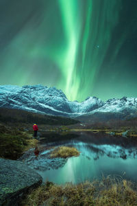 Unrecognizable traveler standing near pond in rocky snowy mountains and admiring view of green aurora borealis illuminating dark night sky in lofoten islands