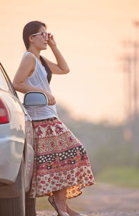 Side view of woman wearing sunglasses standing by car during sunset