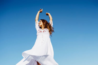 Low angel view of woman with arms raised against clear blue sky