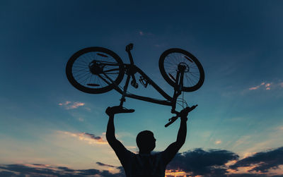 Low angle view of silhouette man holding bicycle against sky