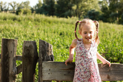 A little girl with pigtails stands against the background of a green field fence made of wood smiles