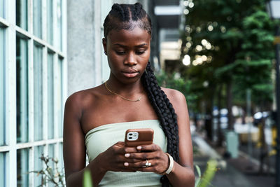 Young african american woman with long braids wearing stylish tube top and browsing smartphone on city street