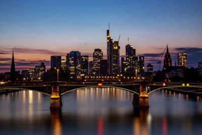 Illuminated bridge over river by buildings against sky in city