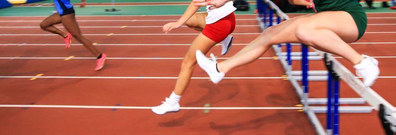 Girls running over hurdles on an indoor track.