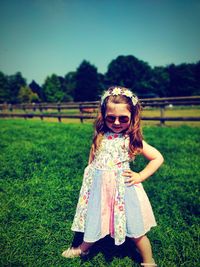 Full length portrait of cute fashionable girl wearing sunglasses and dress standing on grassy field during sunny day