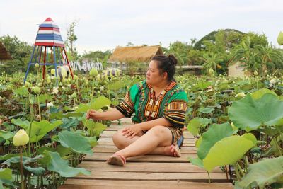 Young woman sitting on boardwalk by flowering plants against sky