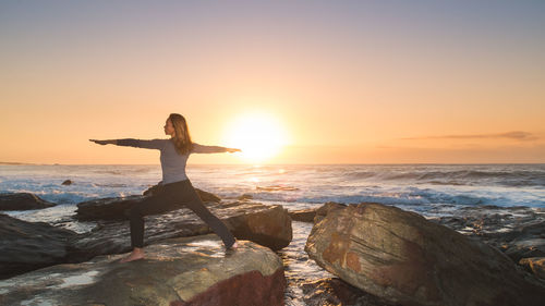 Woman exercising warrior 2 yoga on rock by sea against sky during sunrise