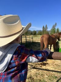 Toddler boy in cowboy hat and flannel hanging on fence looking at horses