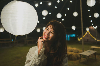 Young woman holding camera while standing against illuminated lights at night