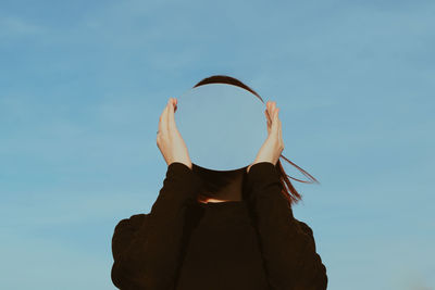 Low angle view of young woman holding mirror against blue sky