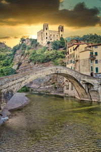 Arch bridge over river by buildings against sky during sunset