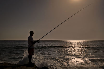 Silhouette man fishing in calm sea at sunset