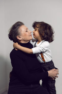 Grandmother with a four-year-old grandson with long hair stands against a white wall with a chair