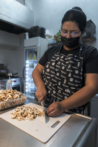 Mature ethnic woman in fabric mask cutting raw mushrooms on chopping board at work in restaurant