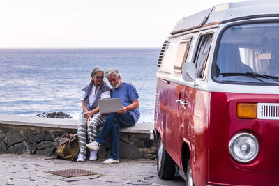 Senior couple traveling in a vintage van, using laptop, sitting on a wall at the sea