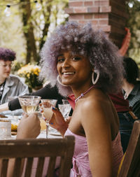 Side view portrait of smiling transwoman with curly hair sitting with friends during party in back yard