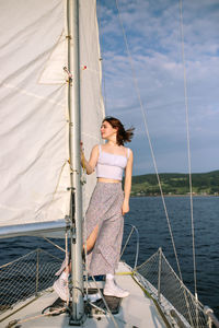 Full body of trendy young female tourist in stylish outfit standing on deck of modern yacht sailing in sea and looking away during summer holidays