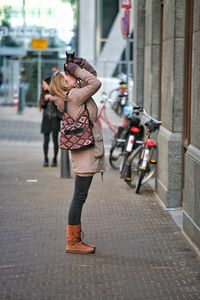 Side view of woman photographing while standing on sidewalk by building