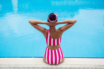Woman in pink striped swimsuit sitting by the pool near the hotel in hot weather