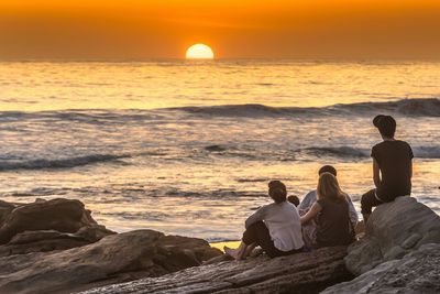 Rear view of friends looking at sunset while sitting on rocks