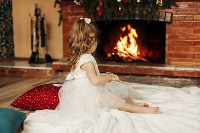 A happy little girl is sitting by the fireplace in a cozy living room on christmas eve