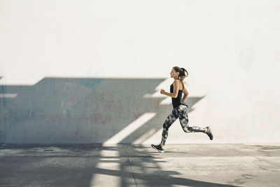Full length side view of female athlete running on sidewalk against white wall
