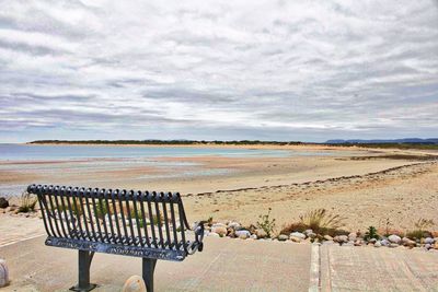 Empty park bench at beach against cloudy sky on sunny day