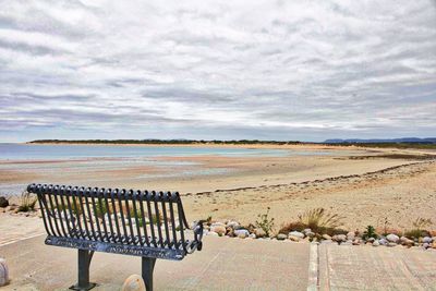 Empty park bench at beach against cloudy sky on sunny day