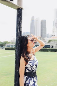 Young woman wearing sunglasses shielding eyes while standing on grassy field in park