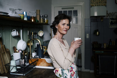Side view of senior woman holding mug while standing in kitchen at home