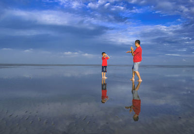 Father playing with son while standing at beach against cloudy sky