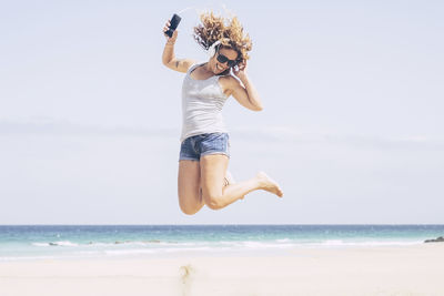 Woman jumping while listening music over phone at beach against sky
