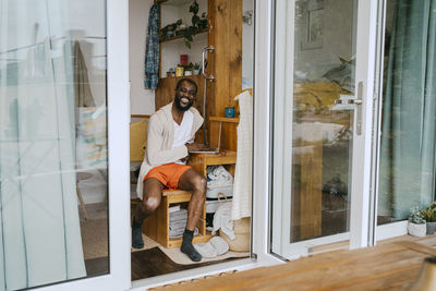 Portrait of happy man with laptop sitting on wooden seat seen through doorway