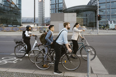 Side view of male and female business professionals waiting with bicycles at roadside in city