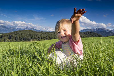 Portrait of smiling cute girl gesturing while sitting on grass against sky