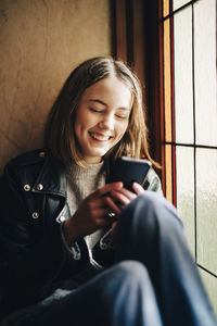 Happy girl using smart phone while sitting by window at home