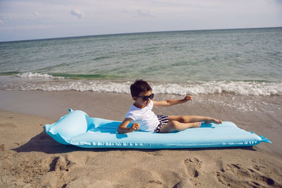 A child boy in sunglasses, a white t-shirt lies on an inflatable mattress on the seashore