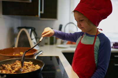 Girl in red chef's hat frying meat in a pan at home