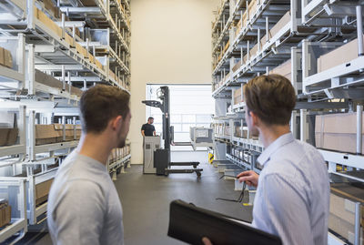 Two men with documents and worker with forklift in factory warehouse