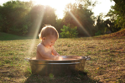 Cute little kid sits in a basin of water in nature and has rays of the setting sun, golden bathe. 