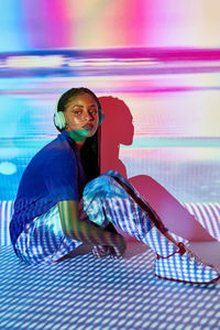 Side view of fashionable young dominican female millennial with long afro braids sitting on floor and looking away while listening to music in headphones in room with colorful geometric illumination