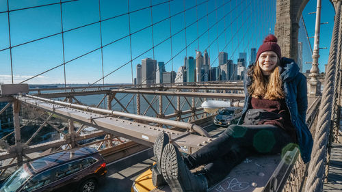 Portrait of smiling woman sitting on bridge against sky in city